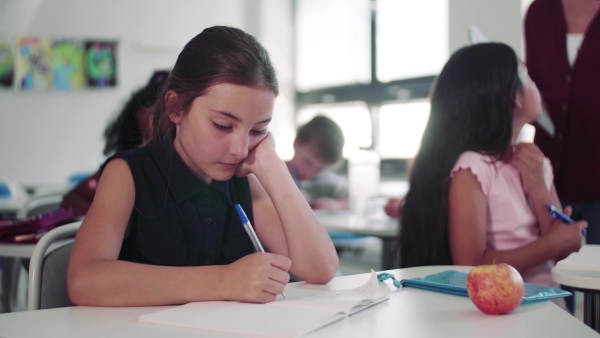 A small bored school girl sitting at the desk in classroom, writing.