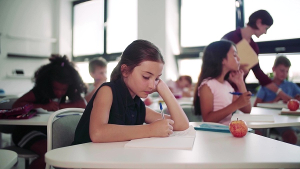 A bored small schoolgirl sitting at the desk on lesson in class, writing. Slow motion.