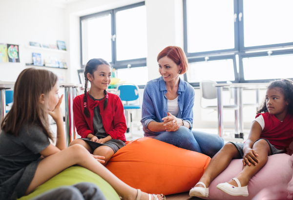 A group of small school kids with teacher sitting on bean bags in class, talking.