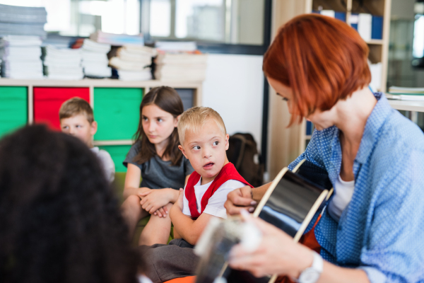 A down-syndrome boy with school kids and teacher sitting on the floor in class, playing guitar.