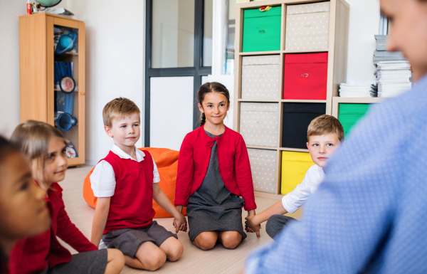 A group of small school kids with unrecognizable teacher sitting on the floor in class, holding hands.