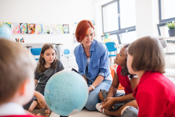 A group of small school kids with unrecognizable teacher sitting on the floor in class, learning geography.