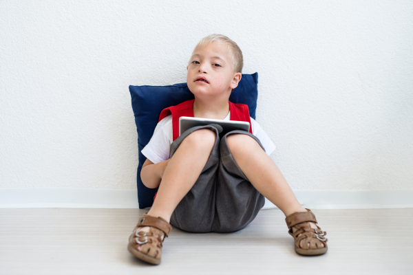 A portrait of happy down-syndrome school boy sitting on the floor, using tablet.