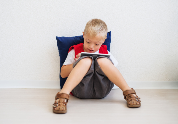 A portrait of happy down-syndrome school boy sitting on the floor, using tablet.