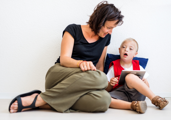 A portrait of down-syndrome school boy sitting on the floor with teacher, using tablet.