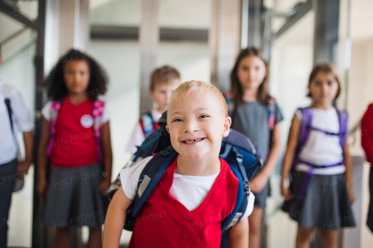 A down-syndrome school boy with bag and group of children in corridor, walking.