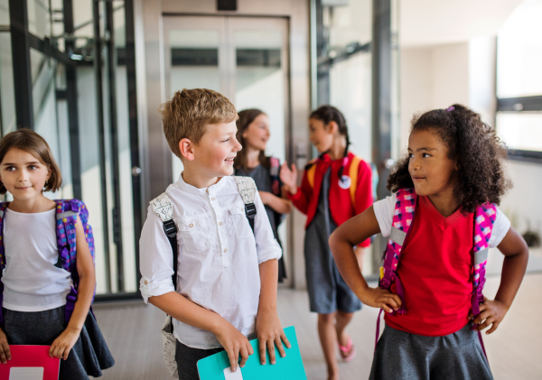 A group of cheerful small school kids with bags walking in corridor, talking. Back to school concept.