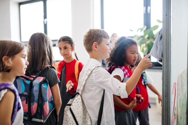 A group of cheerful small school kids with bags standing in corridor, looking at notice board. Back to school concept.