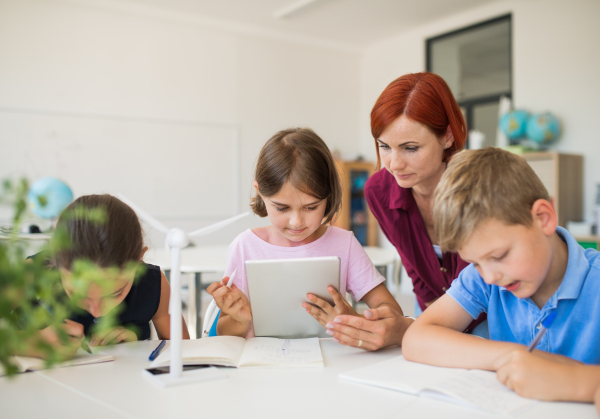 A group of small school kids with teacher sitting in circle in class, using tablet.