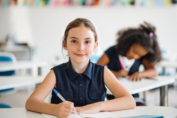 A portrait of small happy school girl sitting at the desk in classroom, looking at camera.