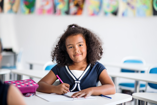 A small mixed race happy school girl sitting at the desk in classroom, writing.