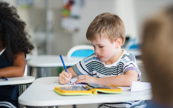 A small happy school boy sitting at the desk in classroom, writing notes.