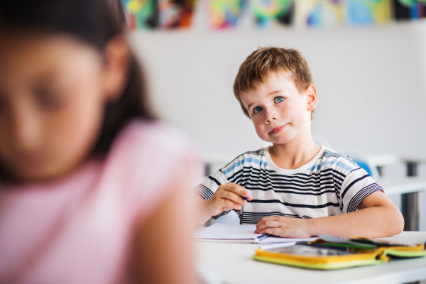 A small happy school boy sitting at the desk in classroom, writing and listening to teacher.