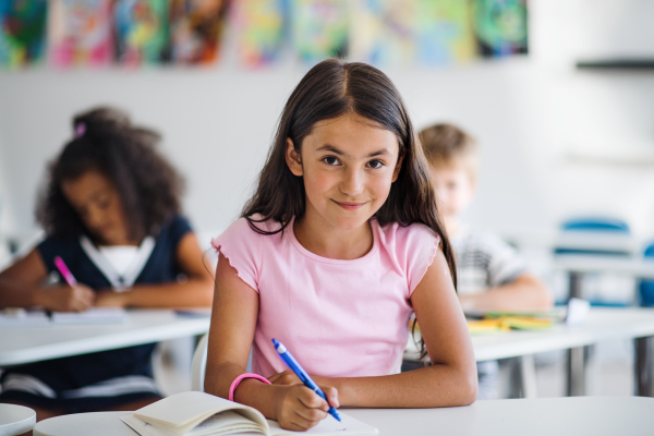 A portrait of small happy school girl sitting at the desk in classroom, looking at camera.