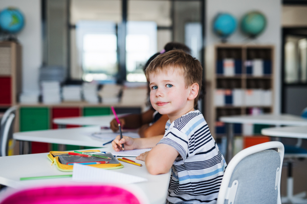 A small happy school boy sitting at the desk in classroom, writing notes.