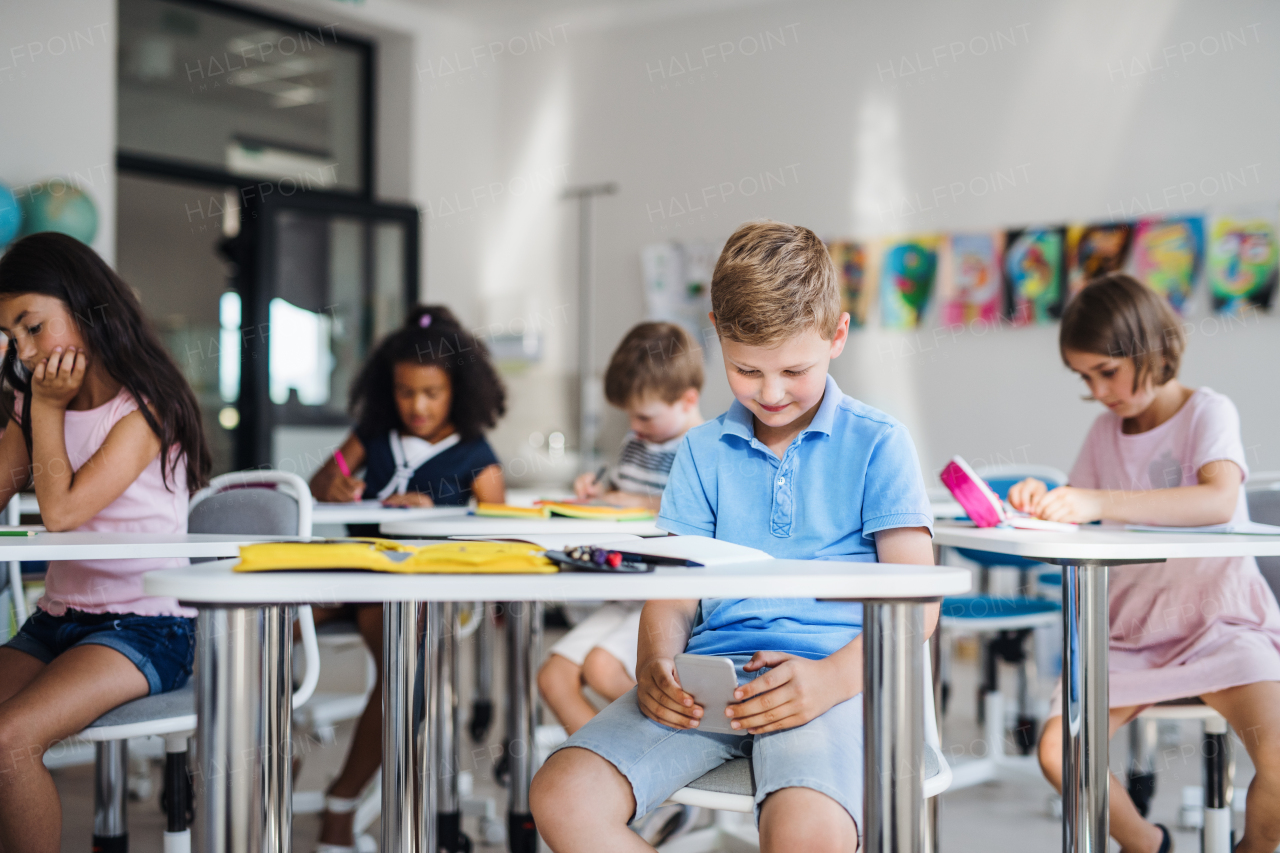 An occupied small school boy with smartphone sitting at the desk in classroom, playing. A danger of addiction.