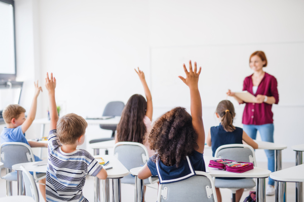 Rear view of school children sitting at the desk in classroom on the lesson, raising hands.