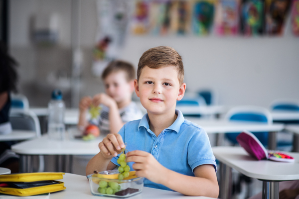 A small happy school boy sitting at the desk in classroom, eating grapes fruit.