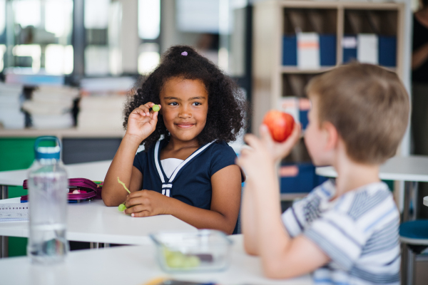 Small school children sitting at the desk in classroom, eating fruit for snack.