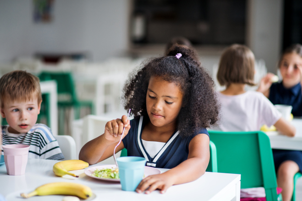 A group of cheerful small school kids in canteen, eating lunch and talking.