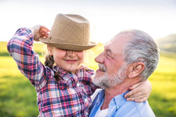 A cheerful small girl with grandfather outside in spring nature, having fun with a hat.