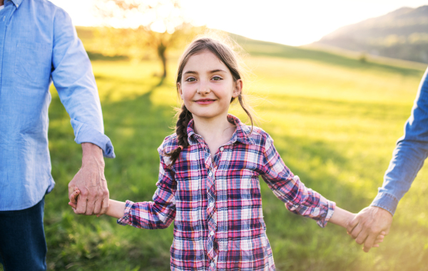 A happy small girl with her unrecognizable senior grandparents walking outside, holding hands. Sunset in spring nature.