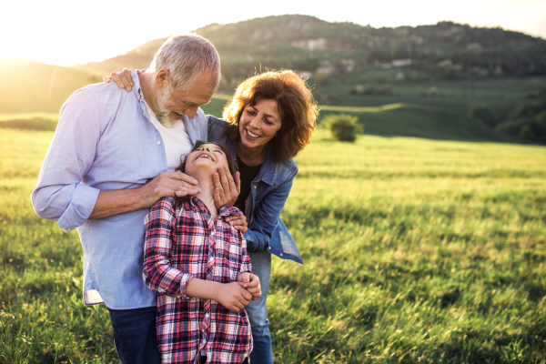 Happy senior couple with granddaughter standing outside in spring nature, looking at each other at sunset. Copy space.