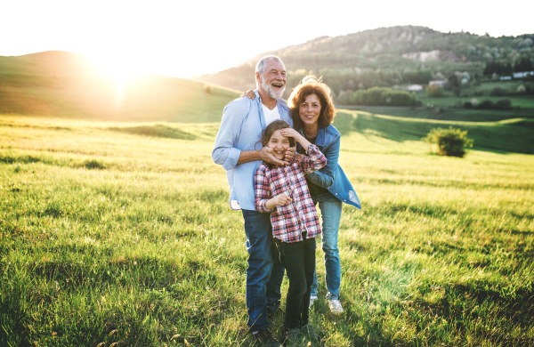 Happy senior couple with granddaughter standing outside in spring nature at sunset.