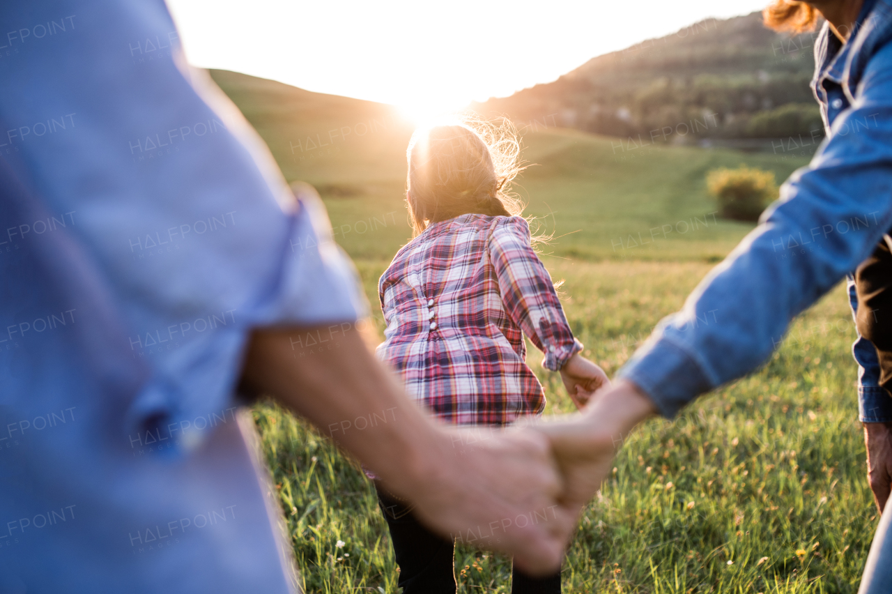 An unrecognizable happy small girl with her senior grandparents having fun outside in nature at sunset. Rear view.