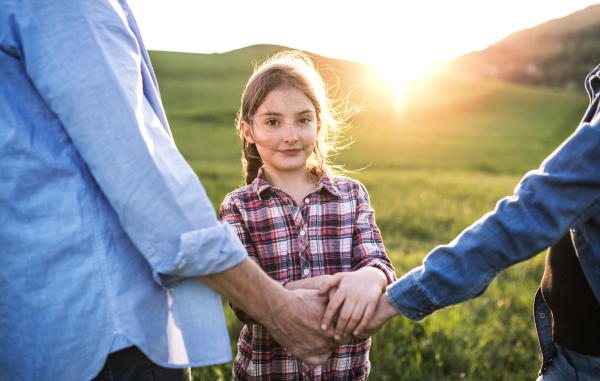 A happy small girl with her unrecognizable senior grandparents standing outside. Sunset in spring nature.