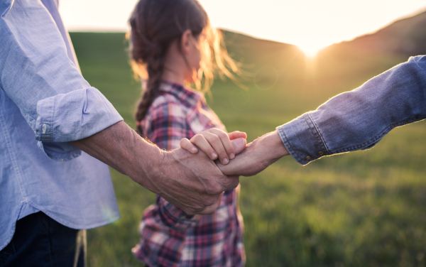 A happy small girl with her unrecognizable senior grandparents walking outside, holding hands. Sunset in spring nature.