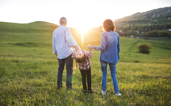 A happy small girl with her unrecognizable senior grandparents having fun outside. Sunset in spring nature. Rear view.