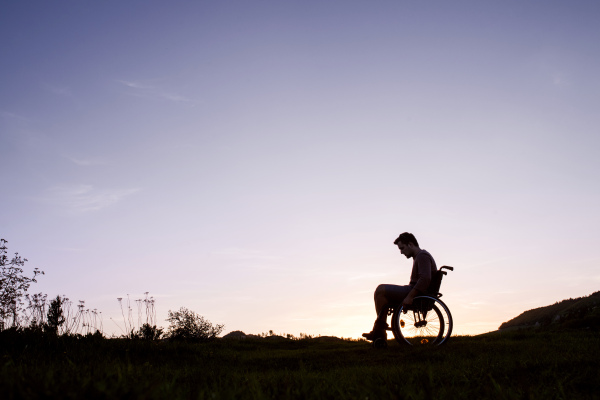 A silhouette of a young man in wheelchair in nature in the evening. Copy space.