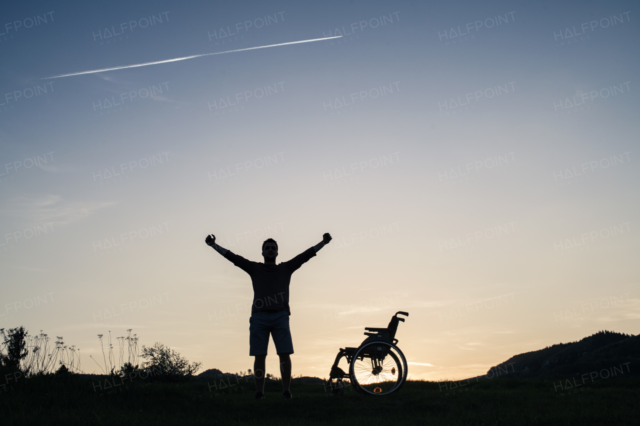 A silhouette of a young man standing by wheelchair in nature in the evening. Copy space.