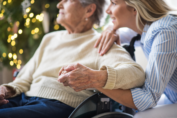 Unrecognizable senior woman in wheelchair with a health visitor at home at Christmas time, looking out of a window.