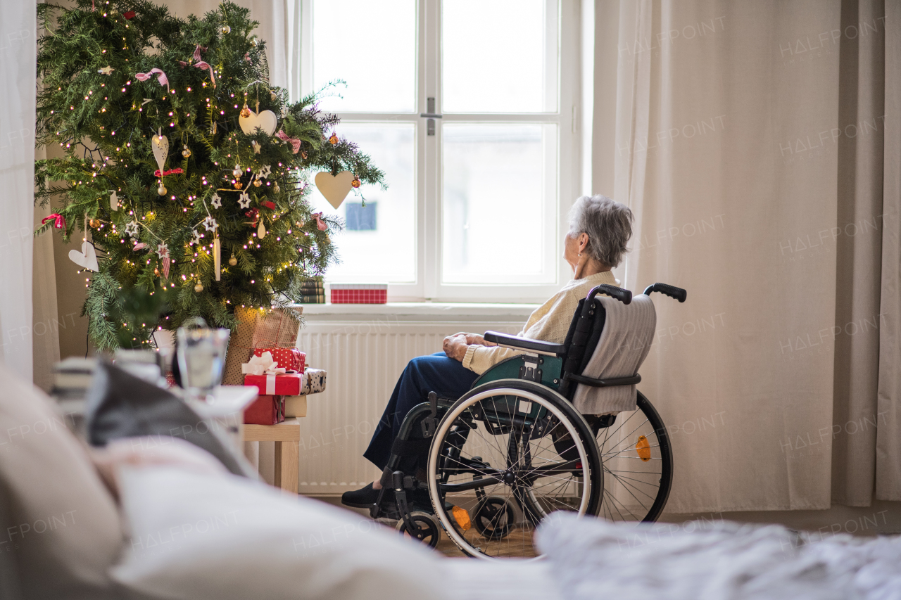 A rear view of a lonely senior woman in wheelchair at home at Christmas time, looking out of a window.