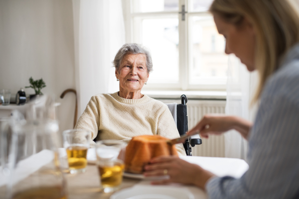 A senior woman in wheelchair with a health visitor sitting at the table at home, cutting a cake.