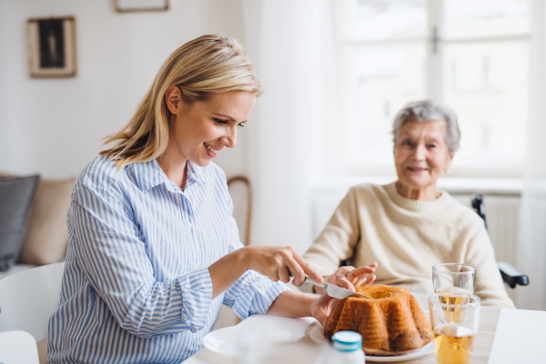 A senior woman in wheelchair with a health visitor sitting at the table at home, cutting a cake.