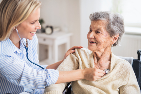 A young health visitor examining a senior woman with a stethoscope at home.