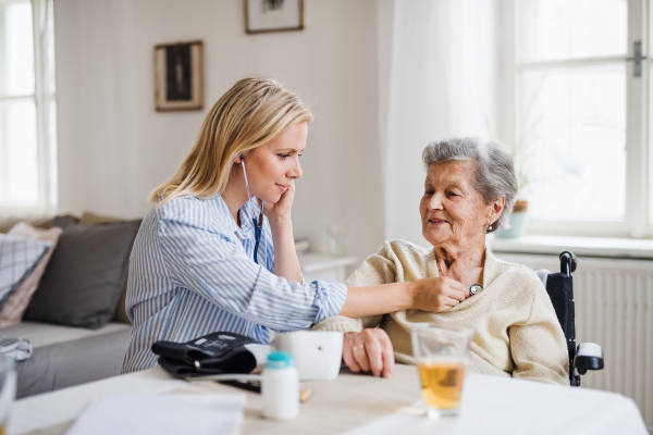 A young health visitor examining a senior woman with a stethoscope at home.