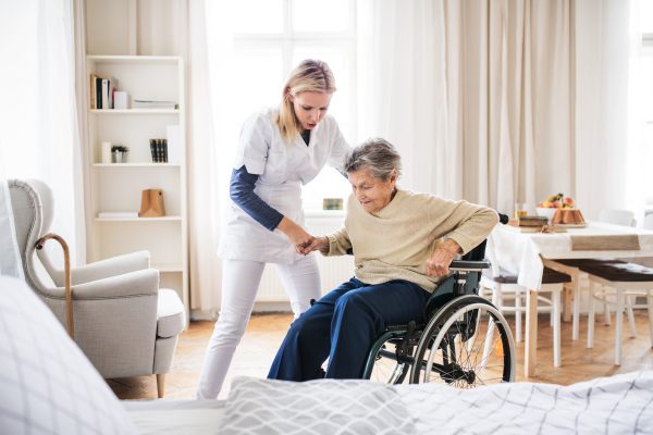 A young health visitor helping a senior woman to stand up from a wheelchair at home.