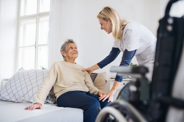 A young health visitor talking to a senior woman sitting on bed at home.