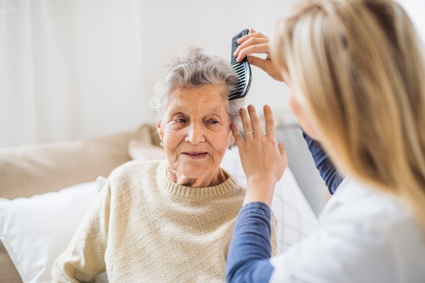 An unrecognizable health visitor combing hair of senior woman sitting on a sofa at home.