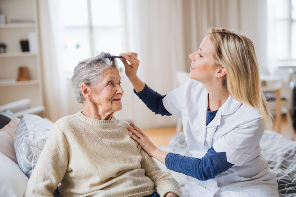 A young health visitor combing hair of senior woman sitting on a bed at home.