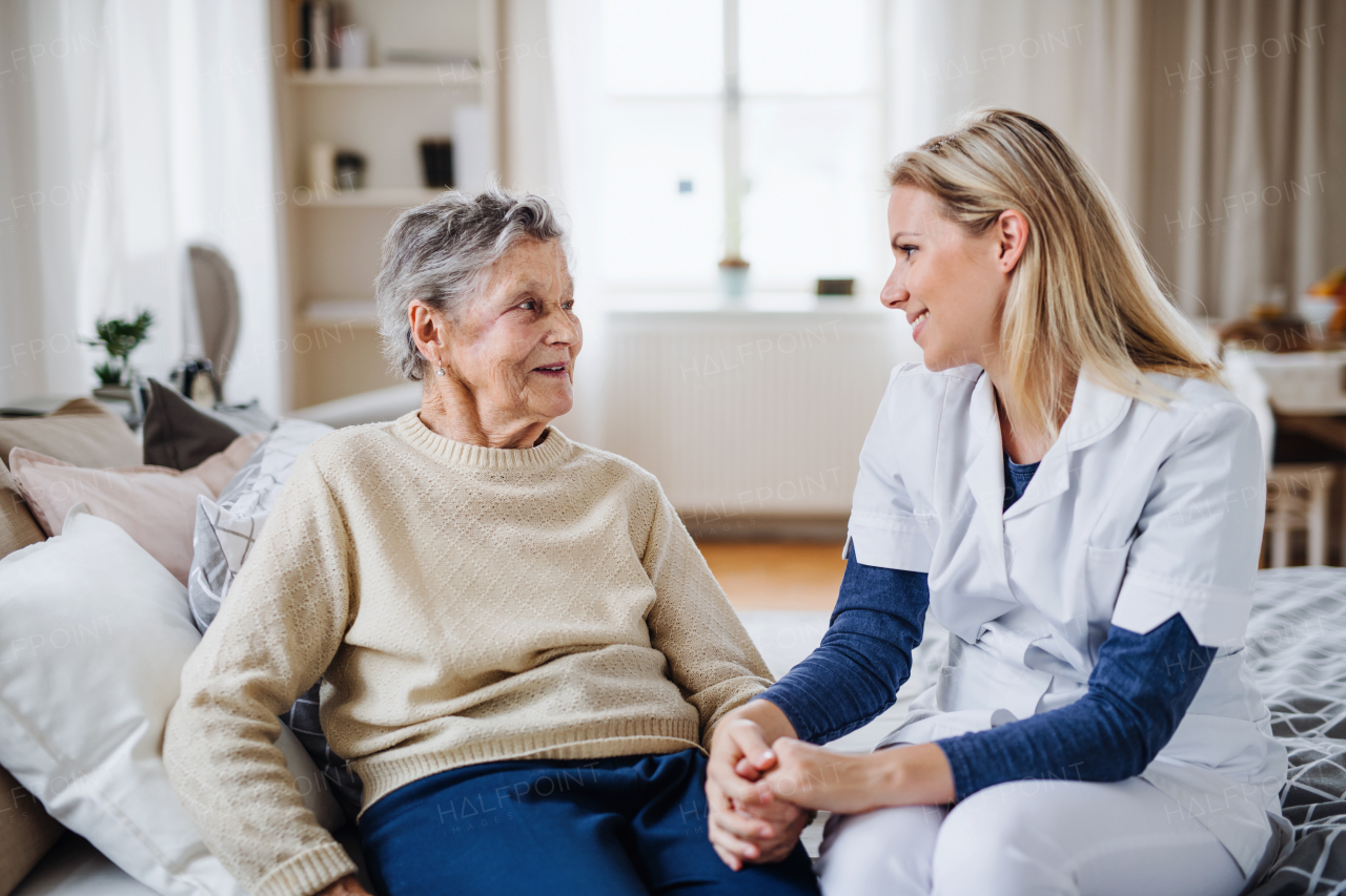 A young health visitor talking to a happy sick senior woman sitting on bed at home, holding her hand.