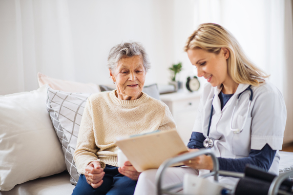 A young health visitor and a senior woman sitting on a bed at home, talking.