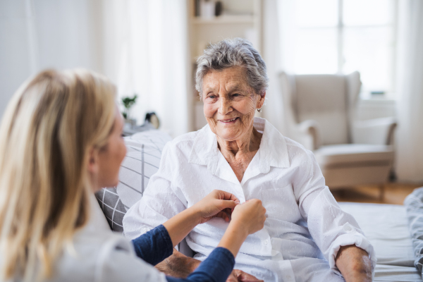 A young health visitor helping a happy sick senior woman sitting on bed at home.