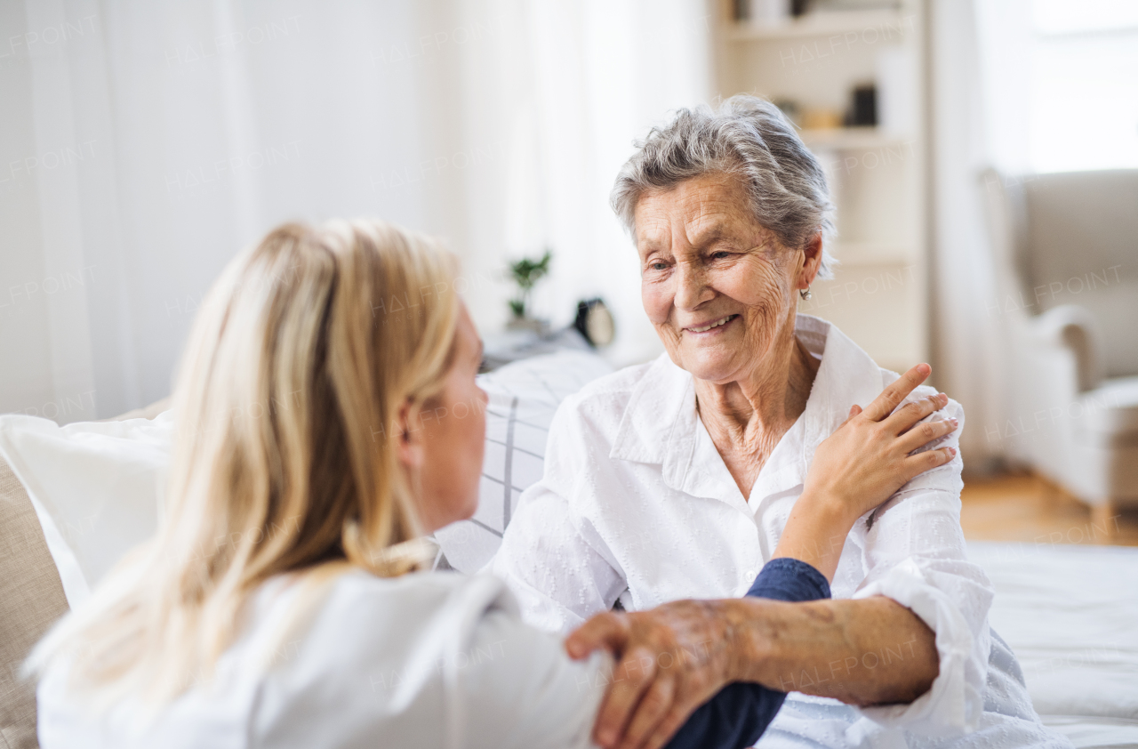 A young health visitor talking to a happy sick senior woman sitting on bed at home.