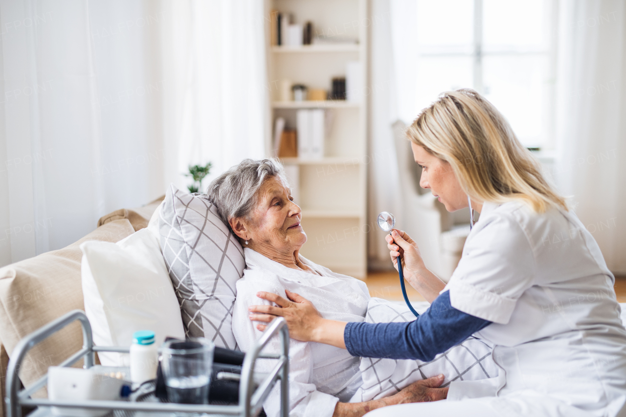 A young health visitor examining a sick senior woman lying in bed at home with stethoscope.