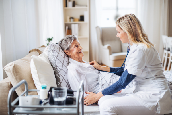 A young health visitor examining a sick senior woman lying in bed at home with stethoscope.